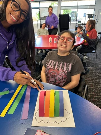 A staff member and participant work on a colorful rainbow paper craft at Cortney's Place, an Arizona charitable tax credit eligible nonprofit day program.