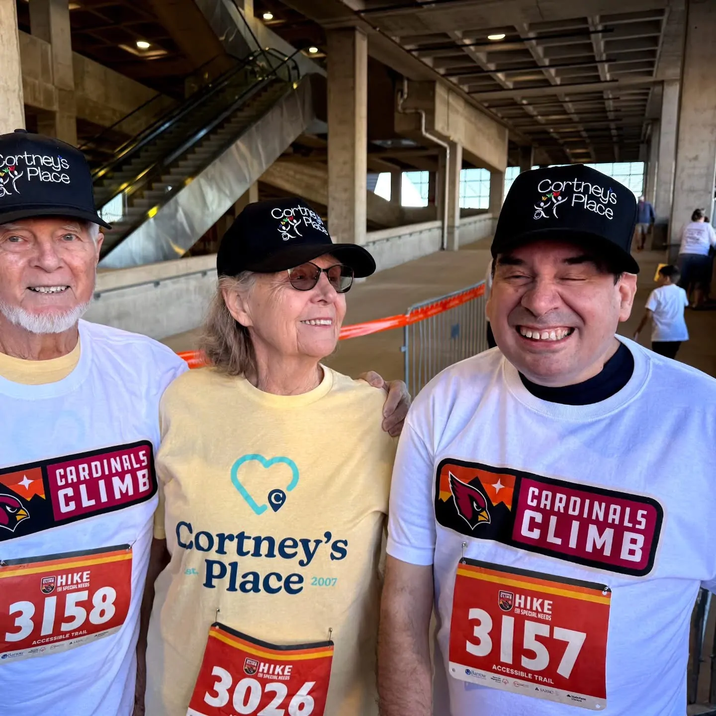 Three smiling Cortney's Place participants wearing Cardinals Climb race bibs and Cortney's Place caps pose together inside State Farm Stadium at the Cardinals Climb fundraising event.