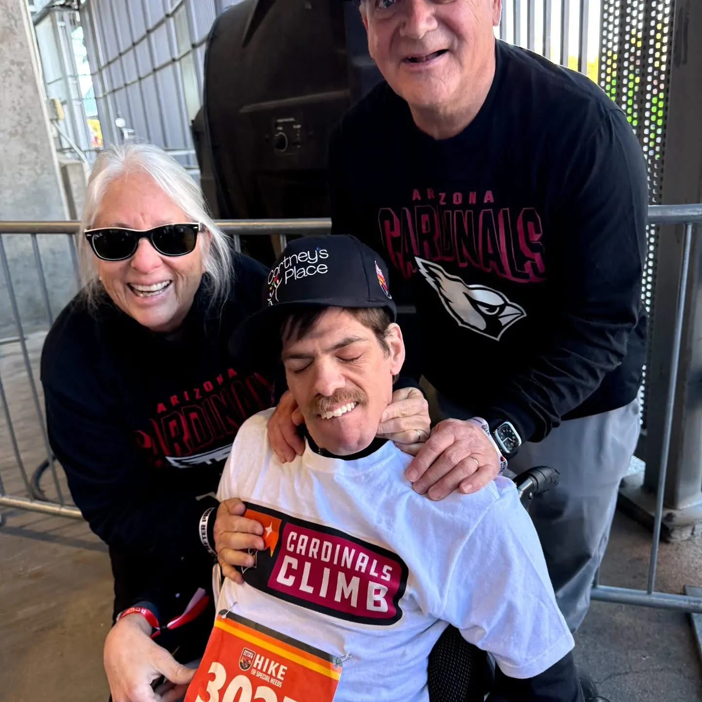 A smiling man in a wheelchair wearing a white Cardinals Climb race bib and a Cortney's Place cap, flanked by two cheerful supporters in Arizona Cardinals long-sleeve shirts at the Cardinals Climb fundraising event.