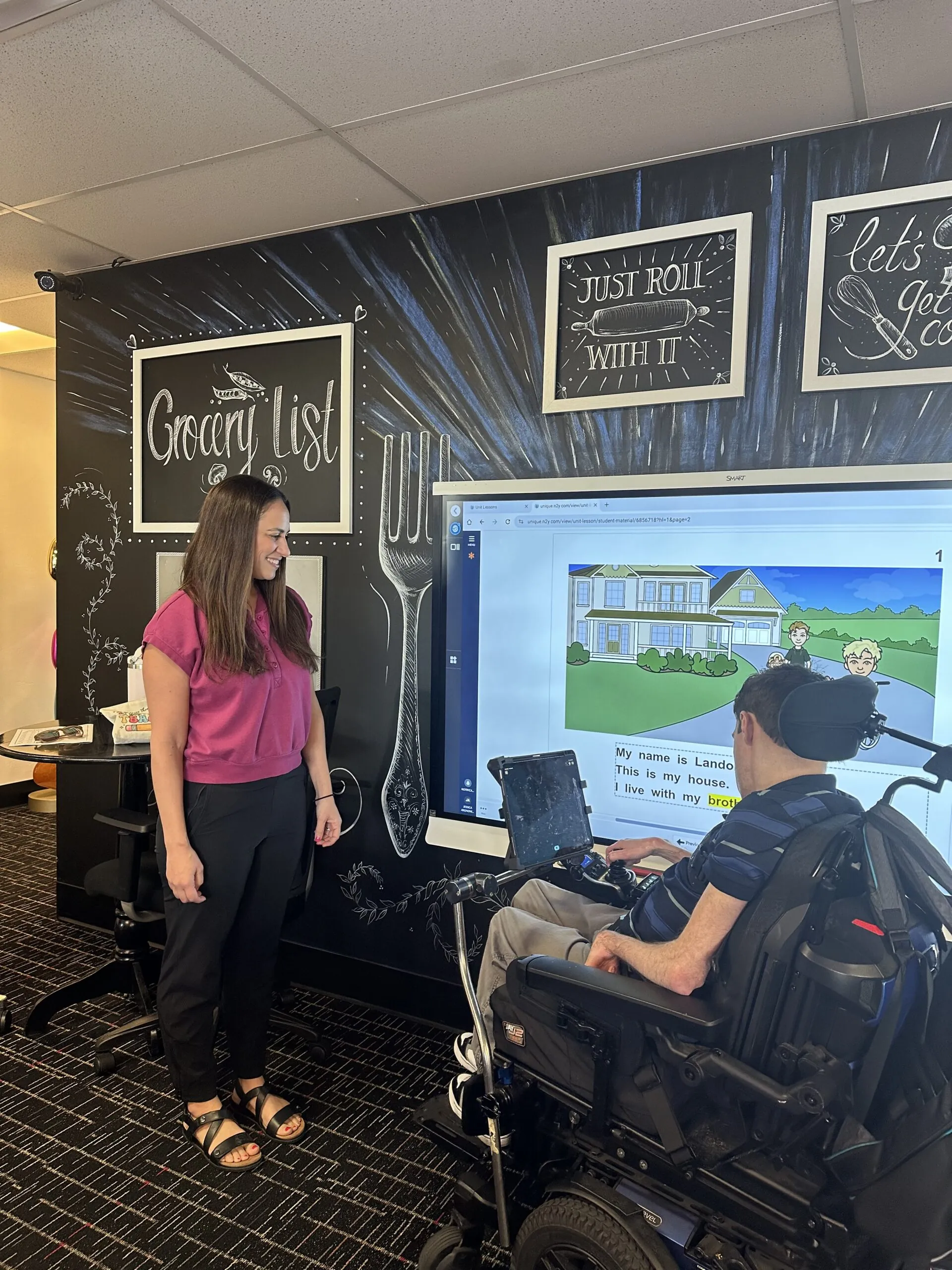 A staff member watches as a participant in a power wheelchair uses a communication device to engage with an interactive literacy lesson on a smart board.