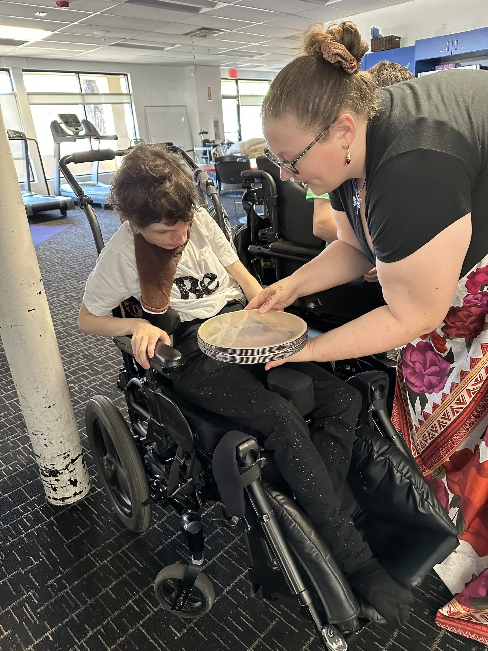 A staff member guides a participant in a wheelchair through a music therapy session using a hand drum inside the day program facility.